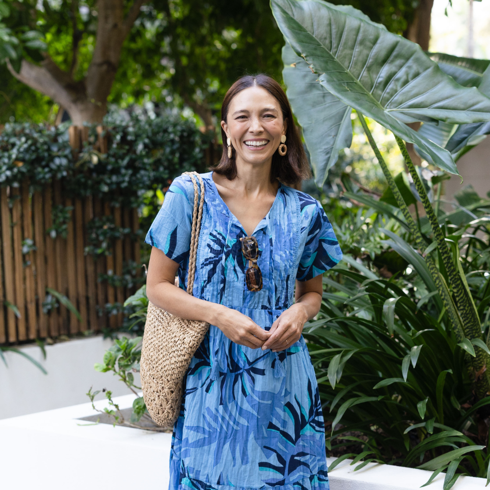 Brunette model wearing a blue floral dress with a woven bag in a tropical garden.