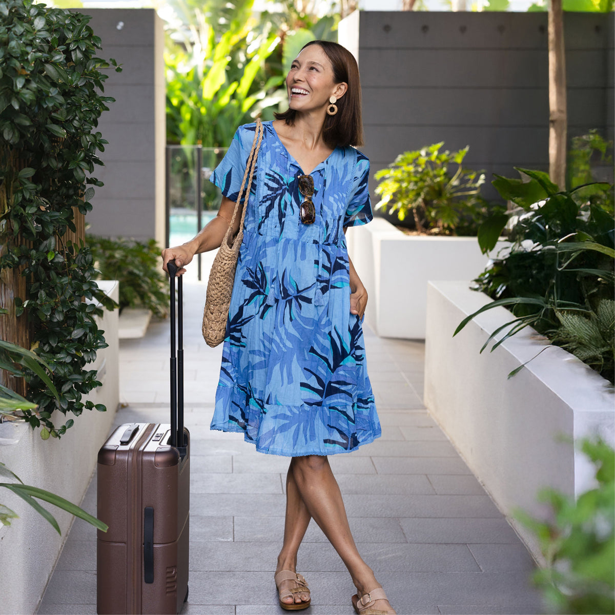 Brunette woman with a suitcase in a loose flowy knee-length blue leaf print sleeved dress, styled with tan sandals.