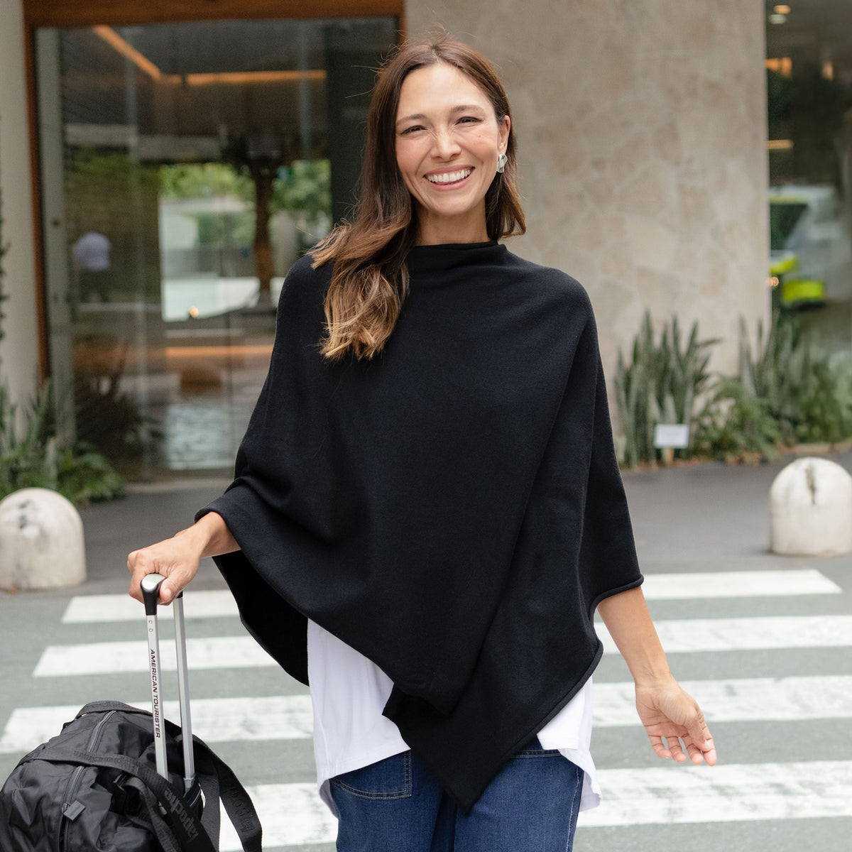 Brunette woman in a black wool blend poncho wheeling a suitcase across the road