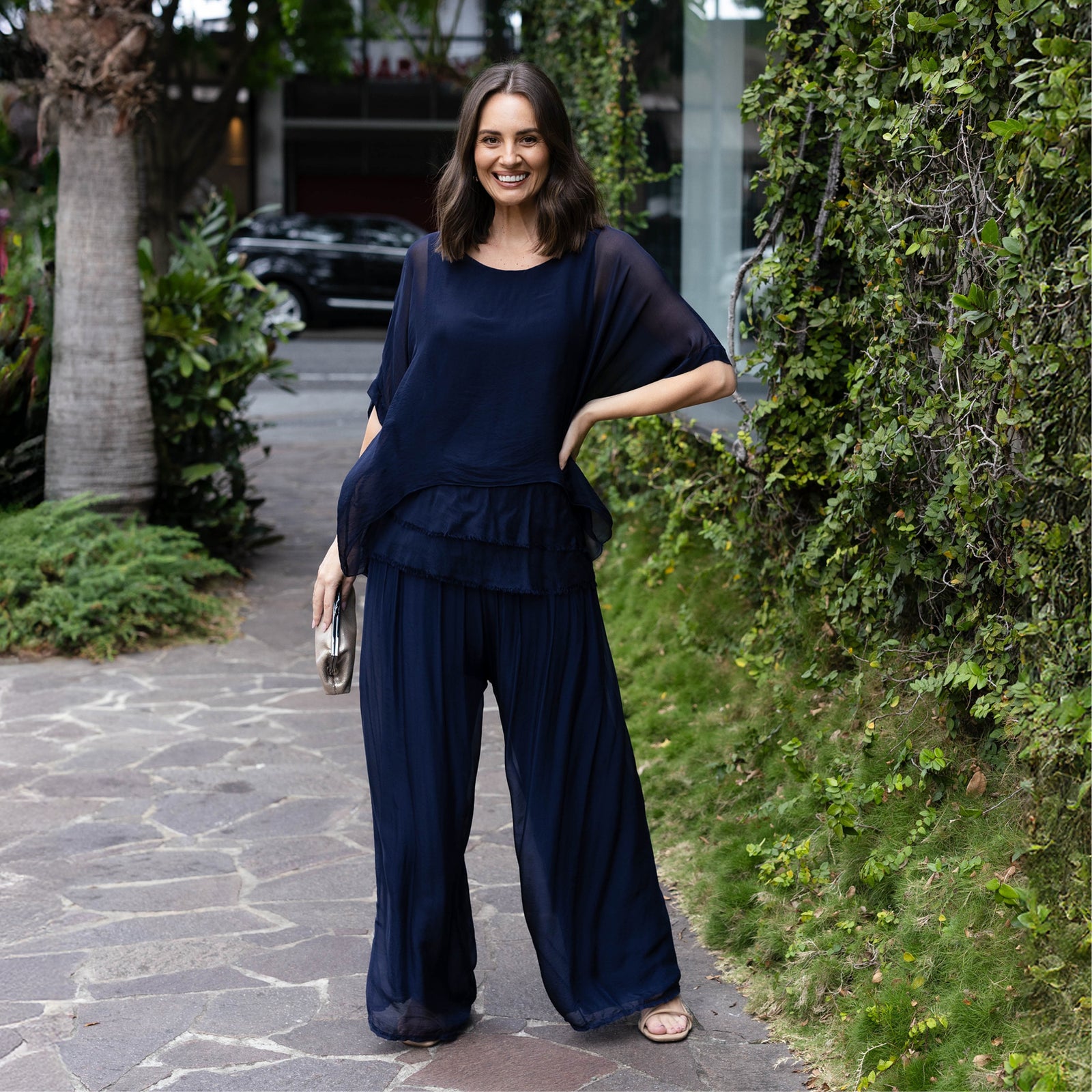 Brunette woman in silk pants and top holding a clutch bag stands with one hand on hip.