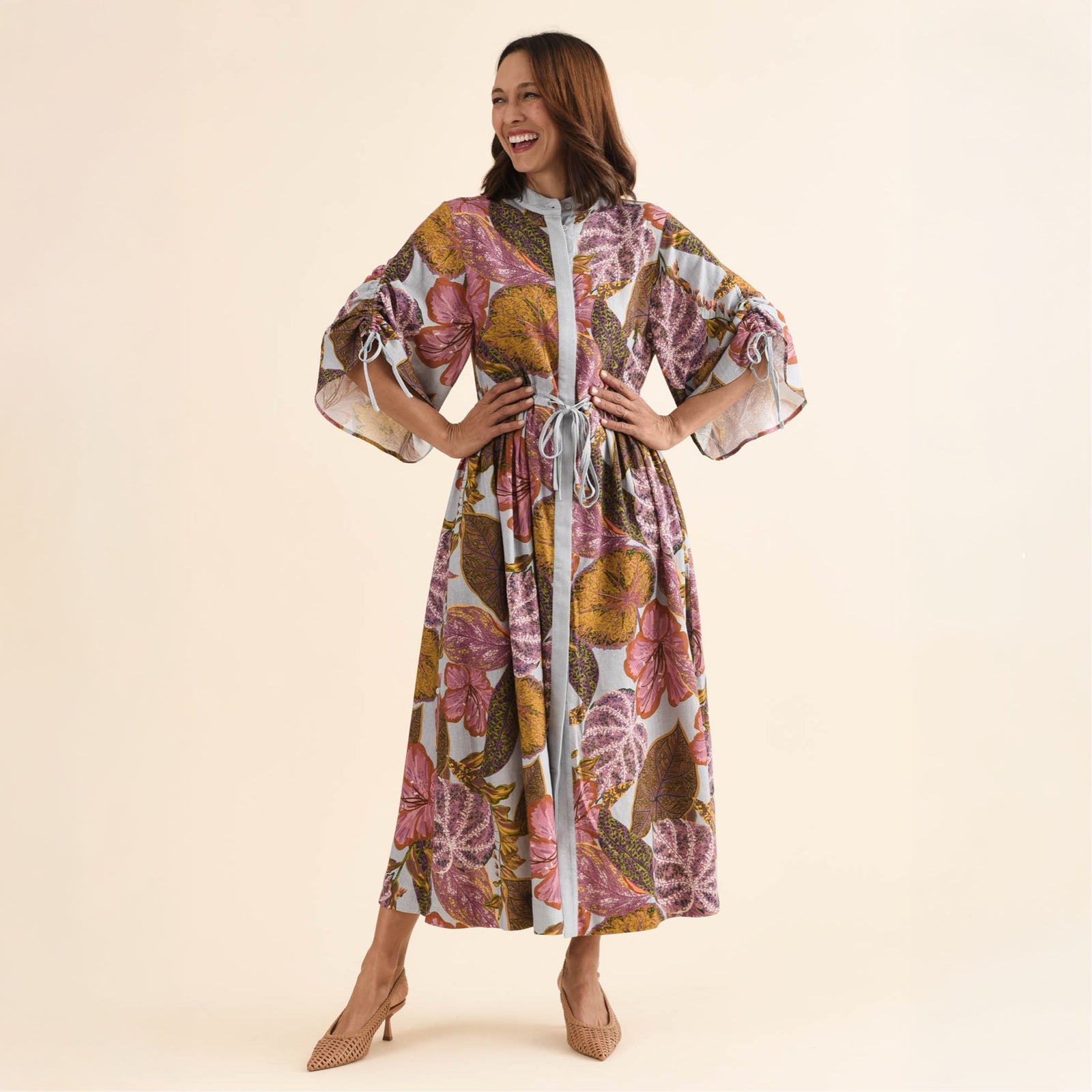 Brunette woman in a printed maxi dress smiles and stands with hands on hips.