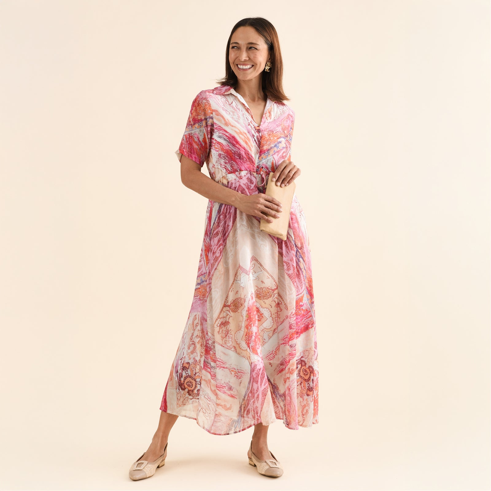 Brunette woman in a printed maxi dress smiles and stands holding a clutch bag.