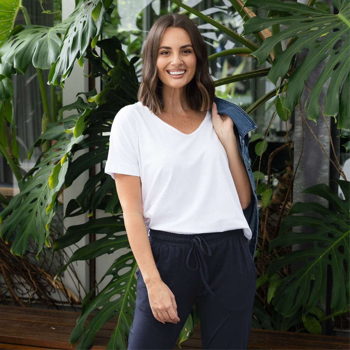 Brunette woman in a white relaxed T-shirt smiles and poses with a jacket over her shoulder.