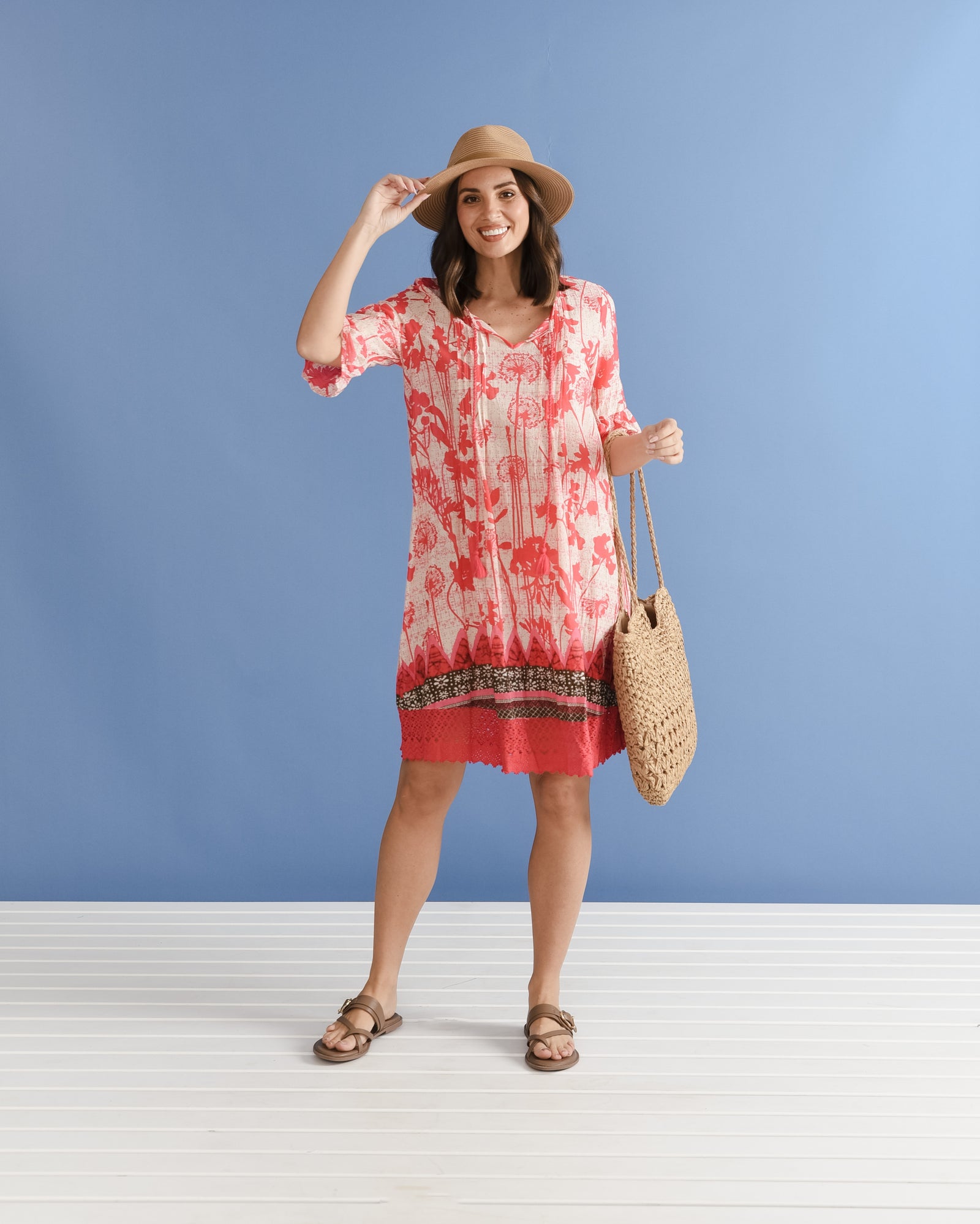 Brunette woman in a hat wearing a coral red pink no iron crinkle cotton dress with brown sandals holding a woven bag.