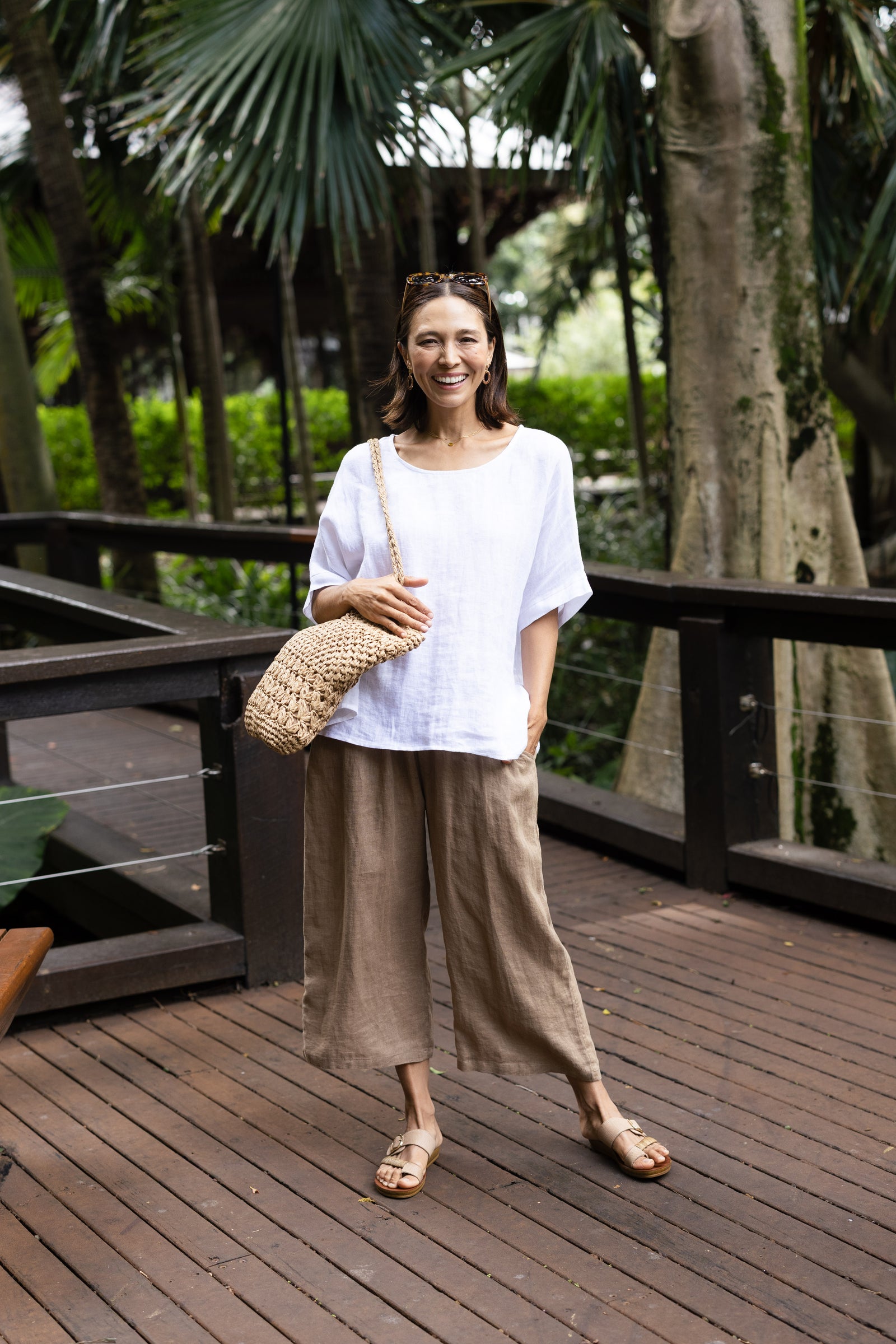 Brunette woman in sunglasses, a white top, light brown pants and sandals with a woven bag.