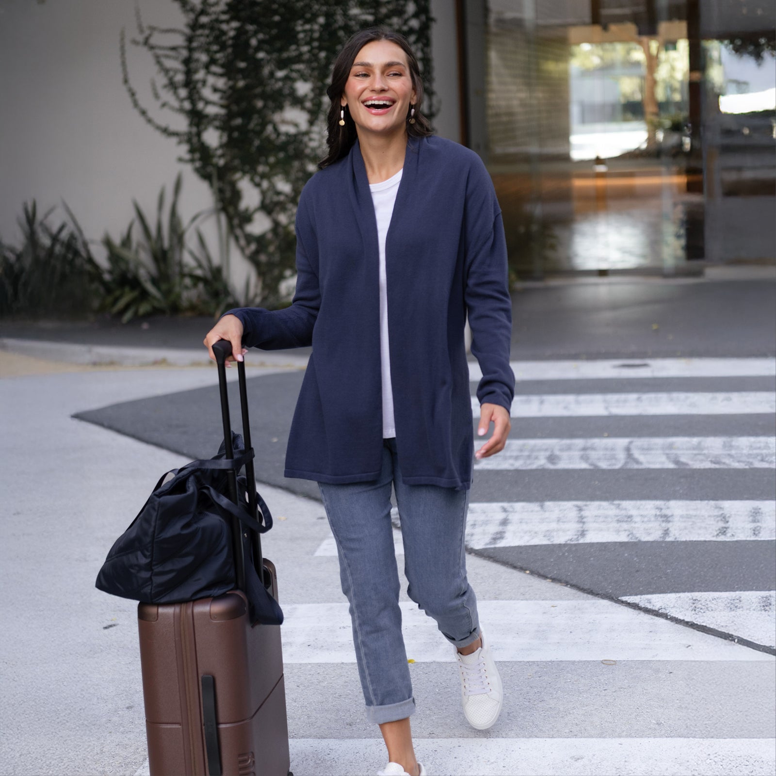 Brunette woman with a suitcase, wearing a cardigan and cropped slim jeans, smiling and walking somewhere.