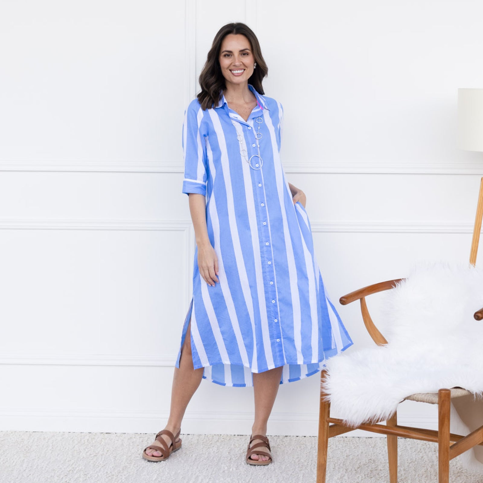 Brunette woman standing in a summery blue and white midi shirt dress and brown flat sandals.