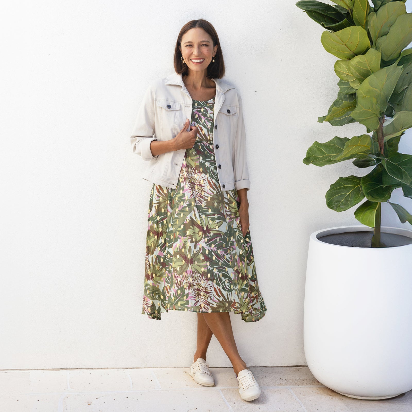 Brunette woman standing in a leafy print green and neutral-toned midi dress with a collared button-up jacket and white sneakers.