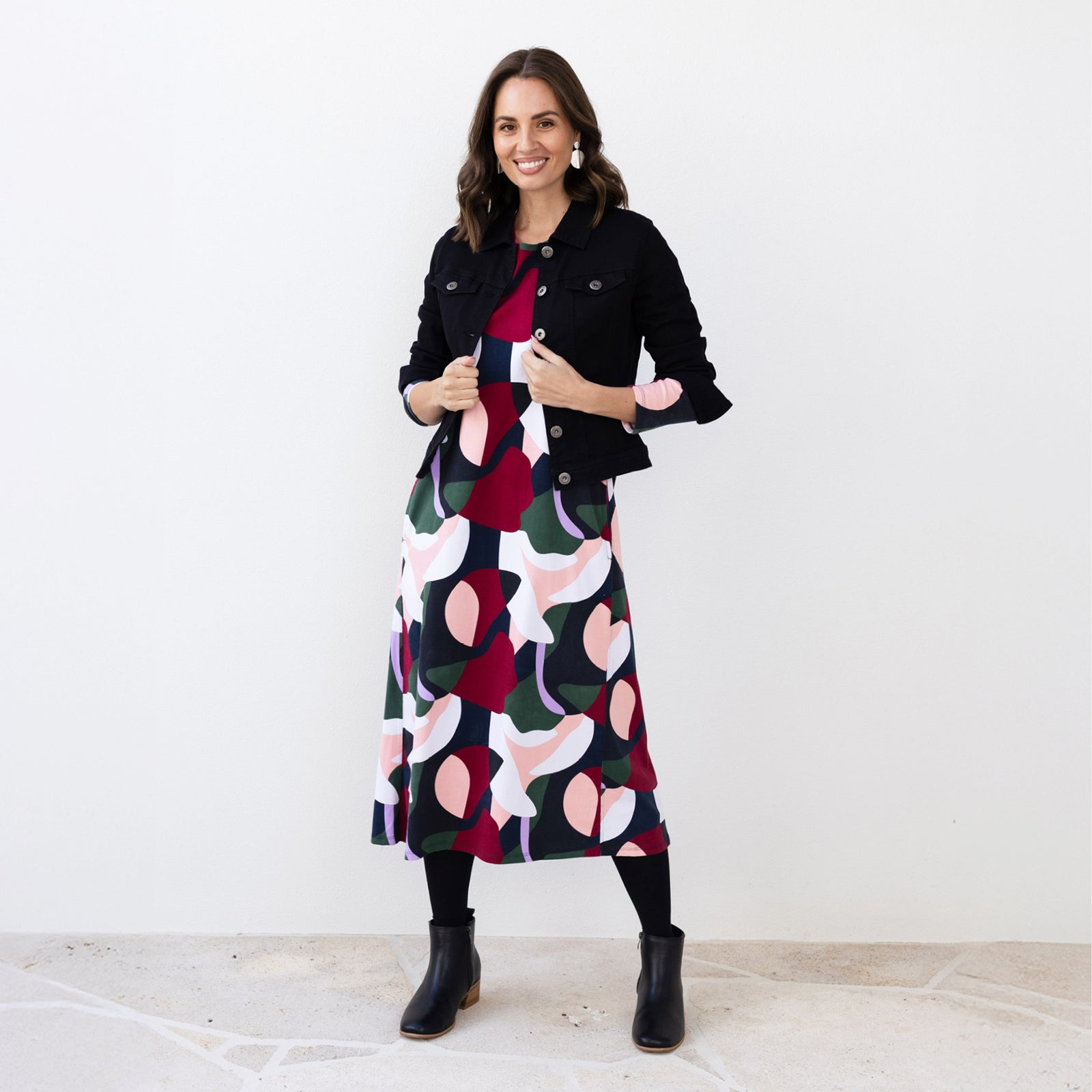 Brunette woman standing in a wintery abstract print red, green, black and white dress styled with boots and a jacket. 
