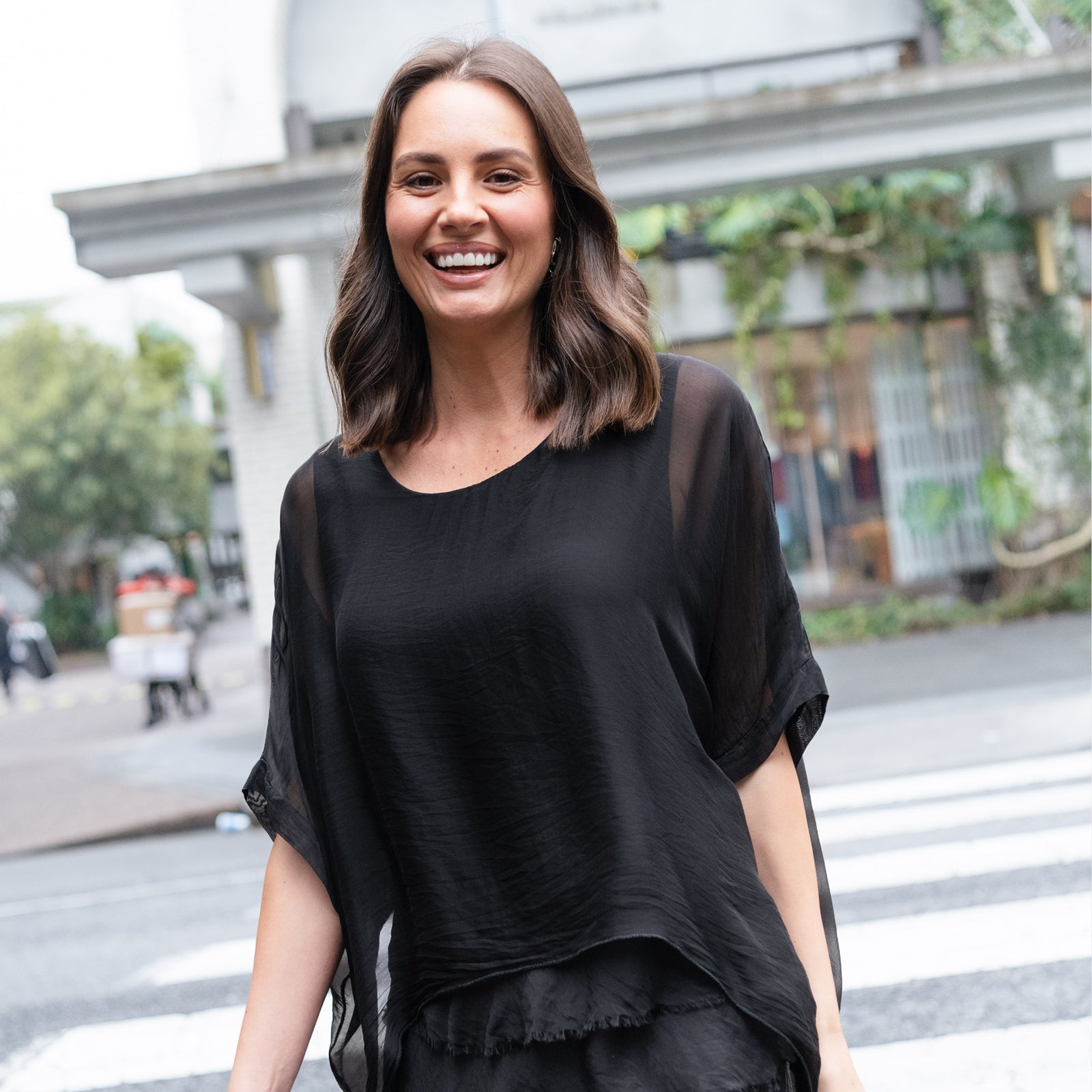 Brunette woman smiles crossing the road in a black wedding guest ensemble.