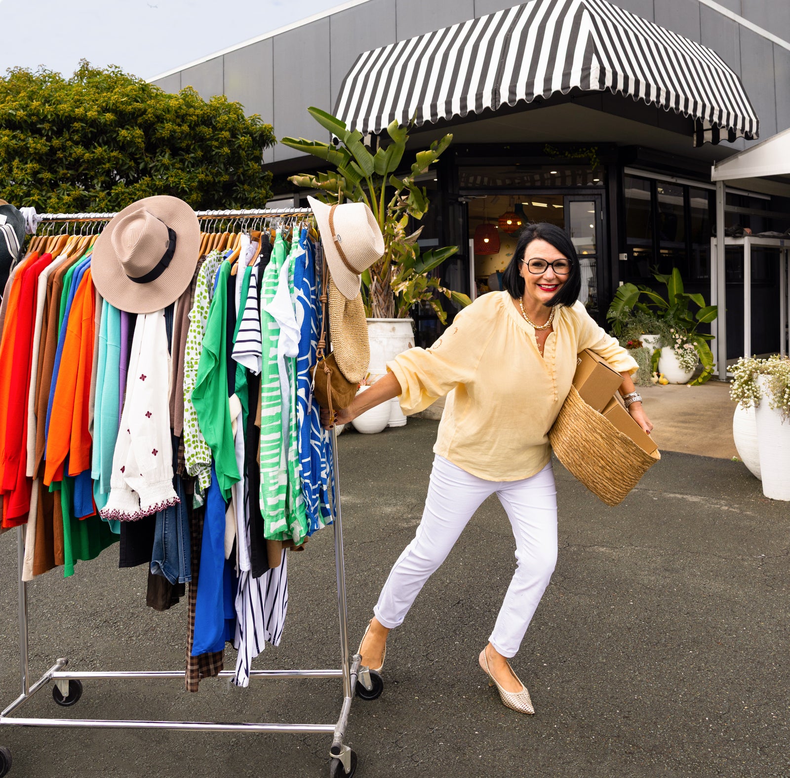 Brunette woman with glasses and red lipstick smiles and pulls a clothes rail across the street. She's wearing white slim pants and a yellow top.