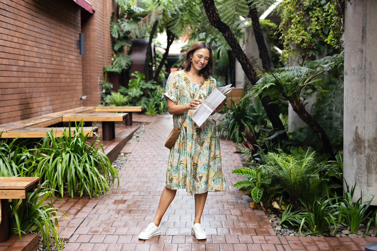 Brunette woman in a blue and green printed dress with white sneakers and a tan cross-body bag reading tourist information.