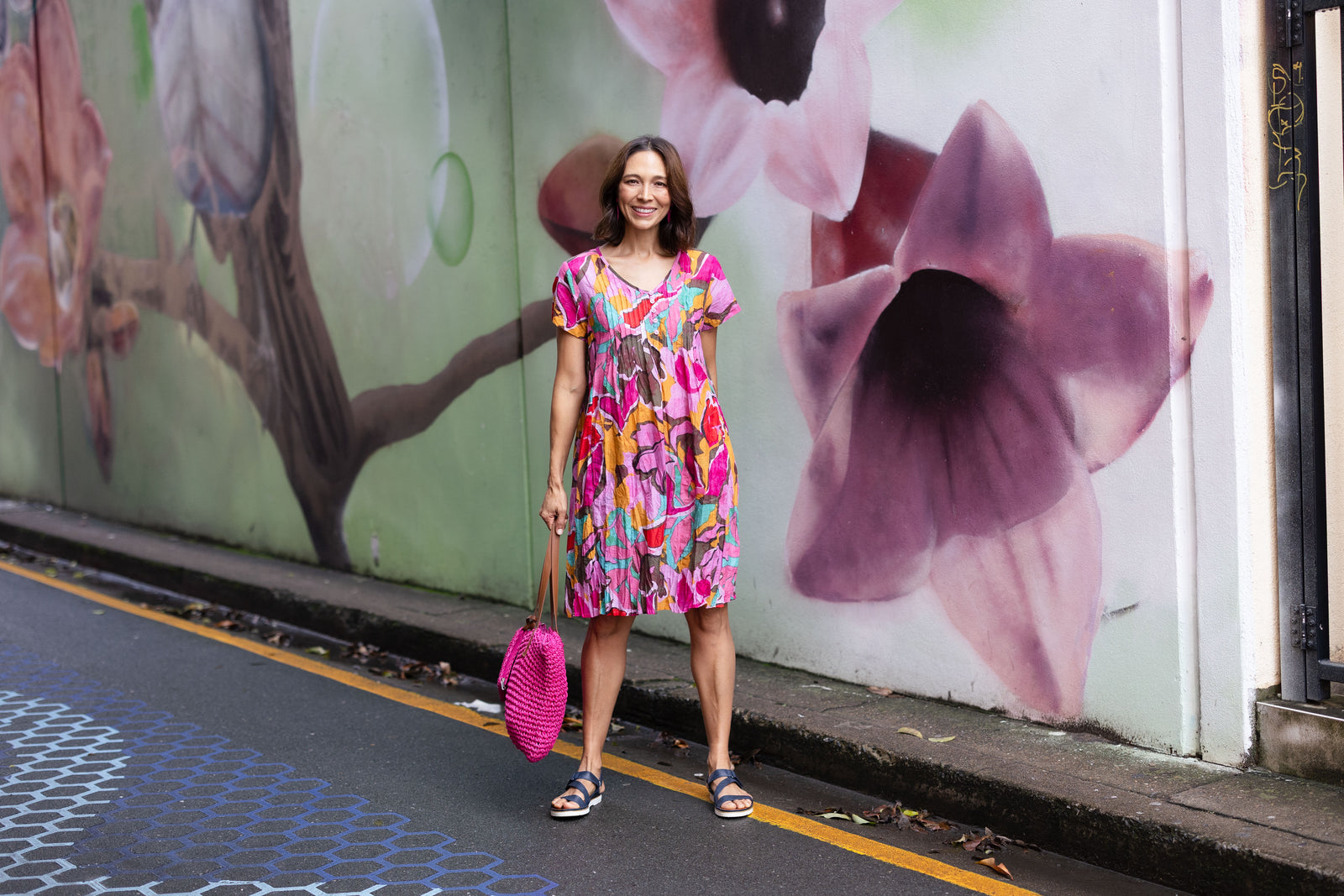 Brunette woman in a bright pink printed no-iron crinkle cotton dress with a matching woven bag and navy sandals stands near a floral mural.
