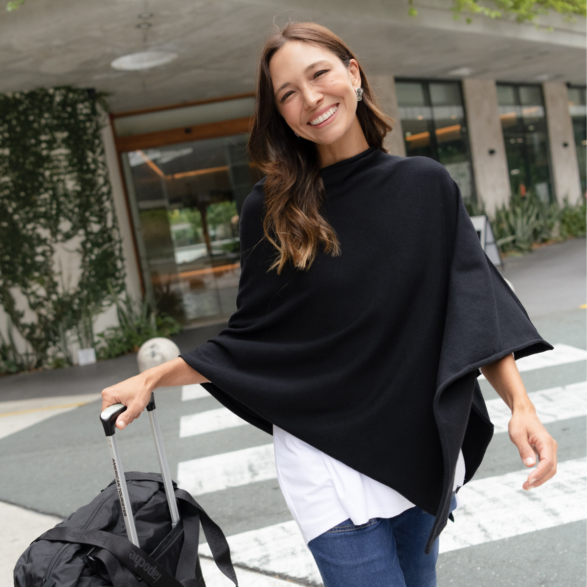 Brunette woman wearing a black poncho wheeling a suitcase.