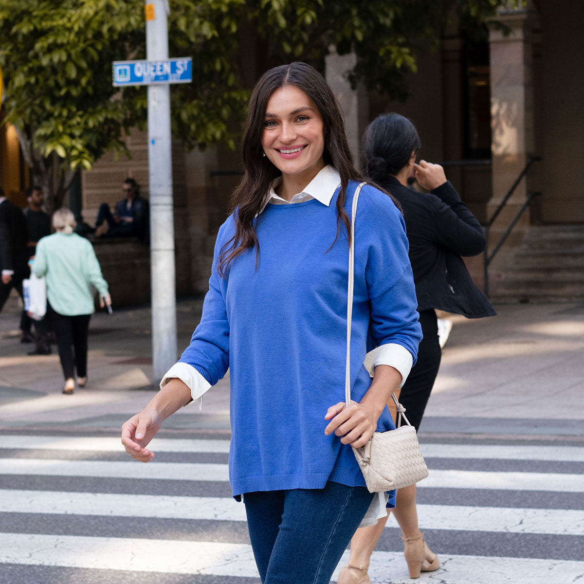 Brunette woman wearing a cobalt blue cardigan walking across the road.